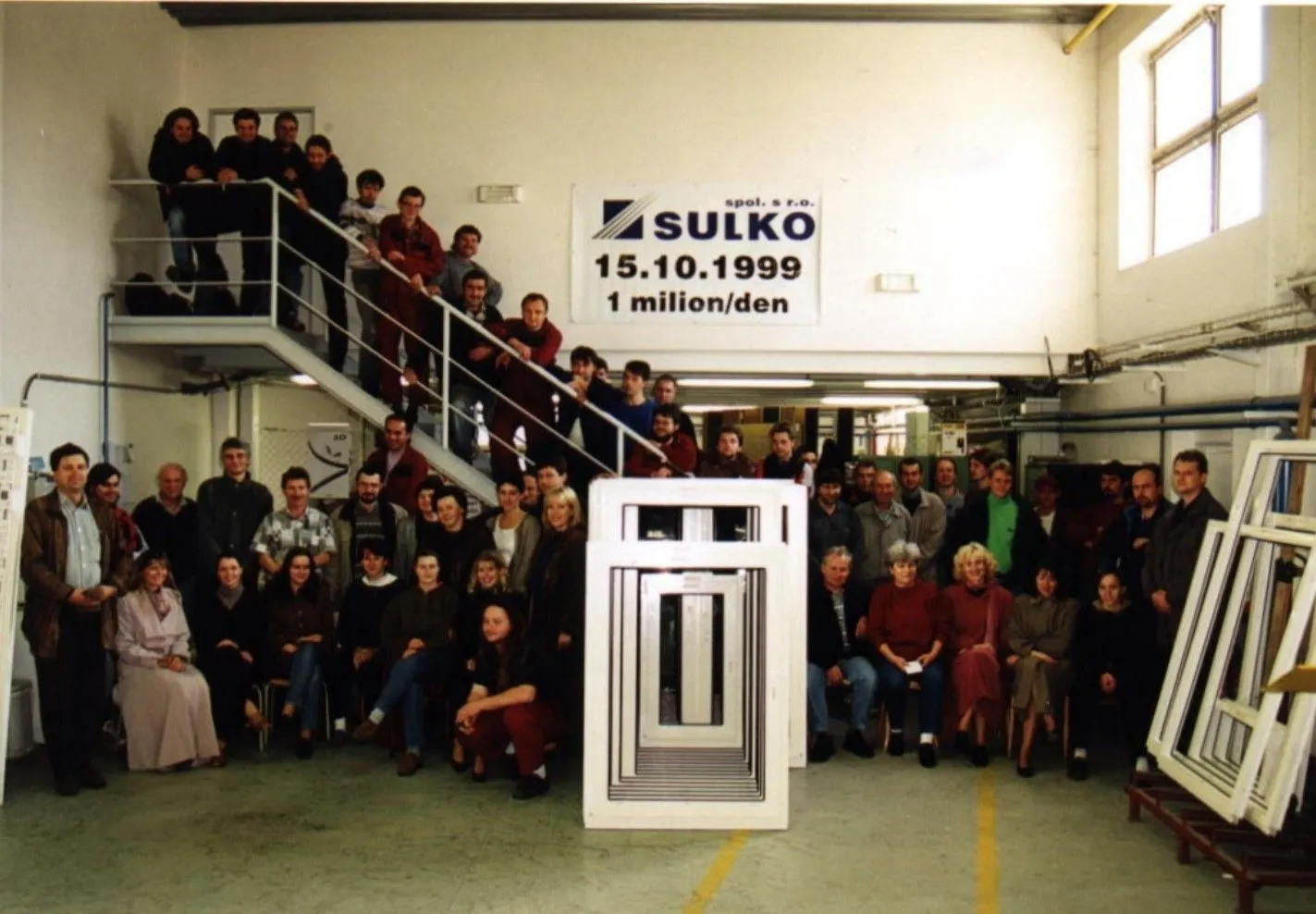 Skupina zaměstnanců stojících a sedících v dílně kolem rámů oken a nápisu Sulko s datem 15.10.1999 a textem 1 milion/den.