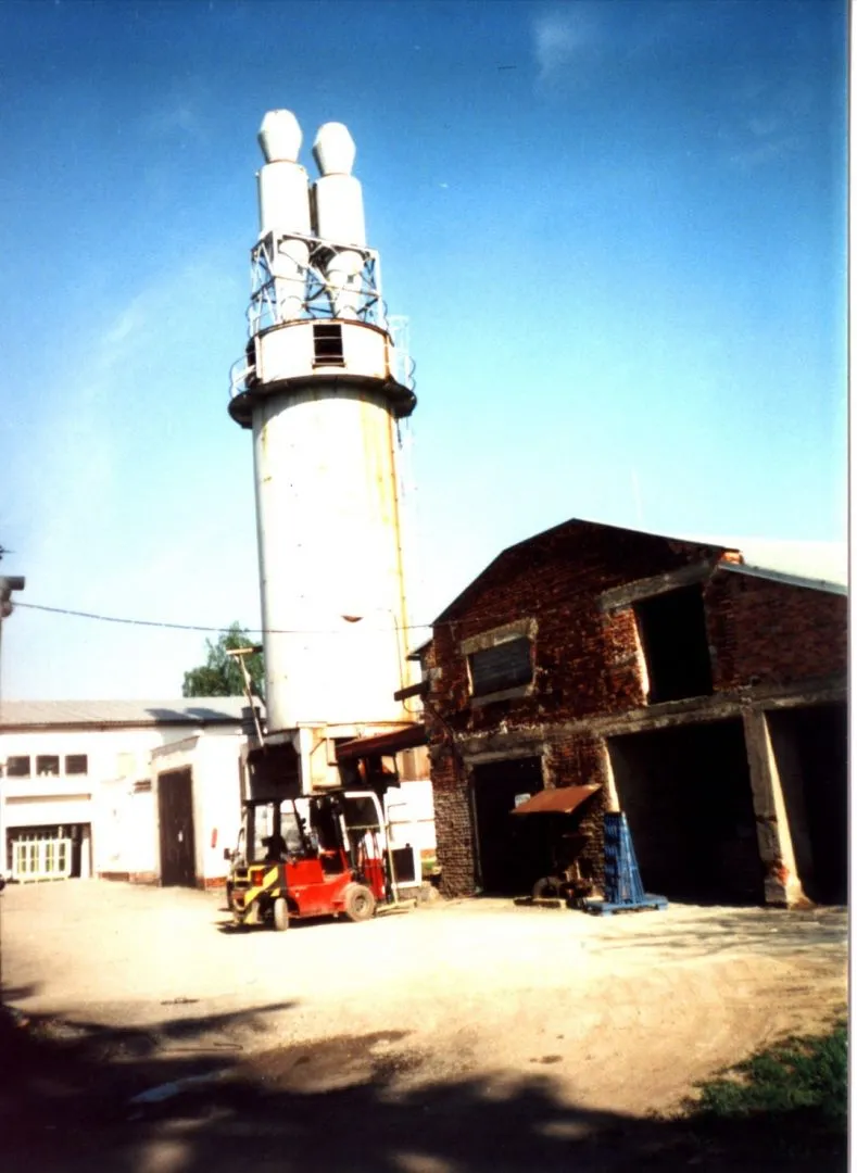 Průmyslový areál s vysokou bílou věží a červeným vysokozdvižným vozíkem vedle staré cihlové budovy za slunného dne.