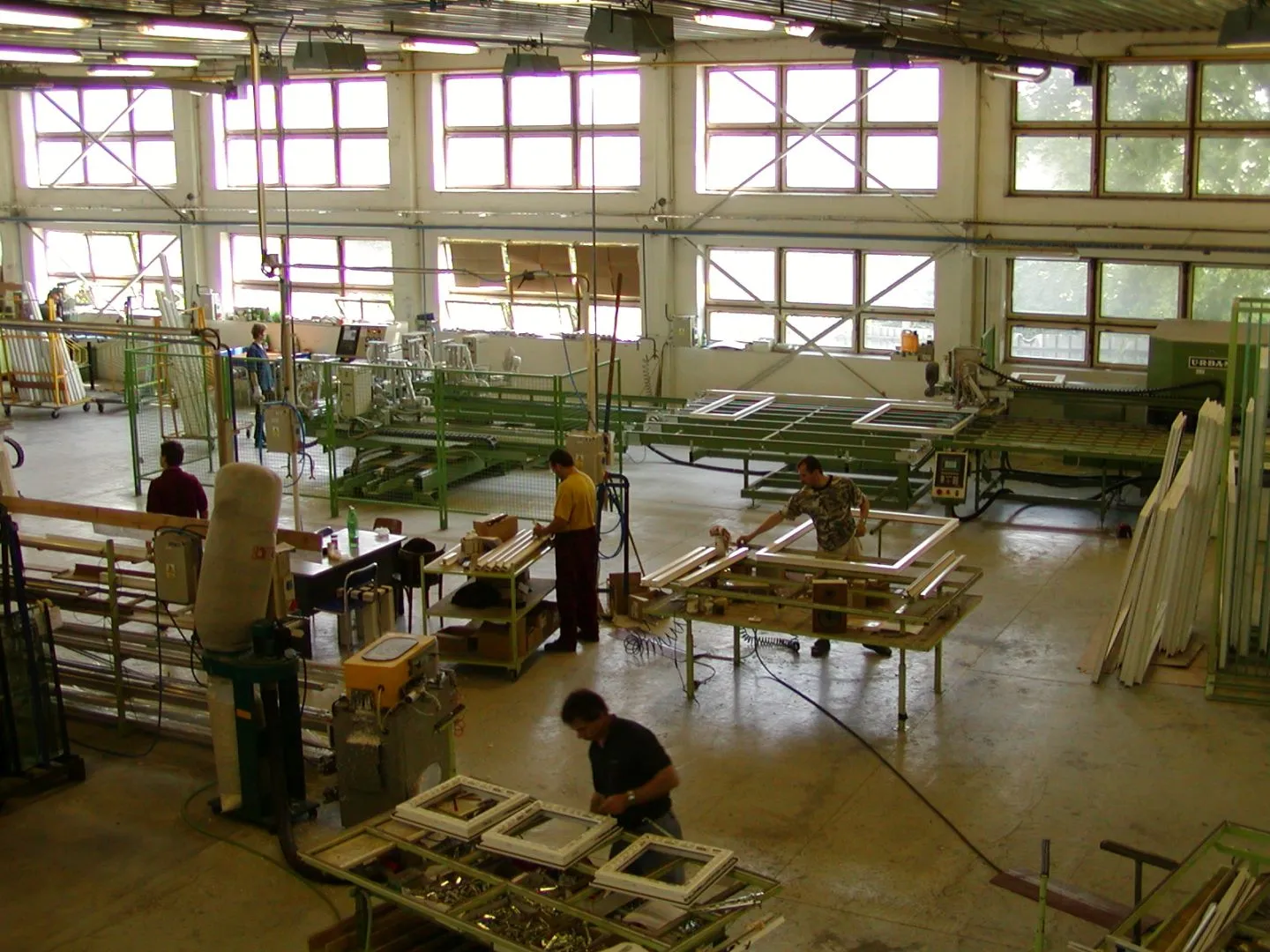 Pracovníci ve výrobní hale sestavují a upravují rámy oken na montážních stolech.