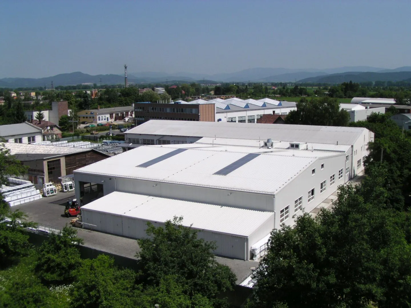 Průmyslový areál s bílými výrobními halami obklopený zelení za jasného dne.