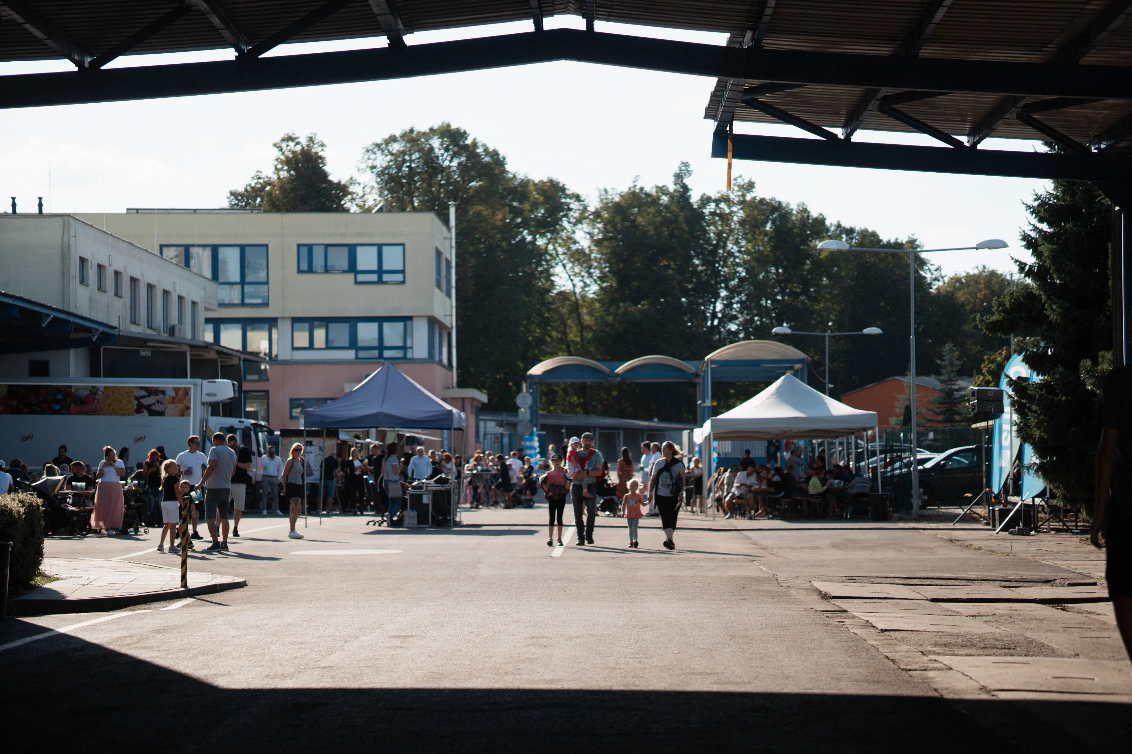 Lidé chodí a sedí pod stanem v průmyslovém areálu za slunečného dne.