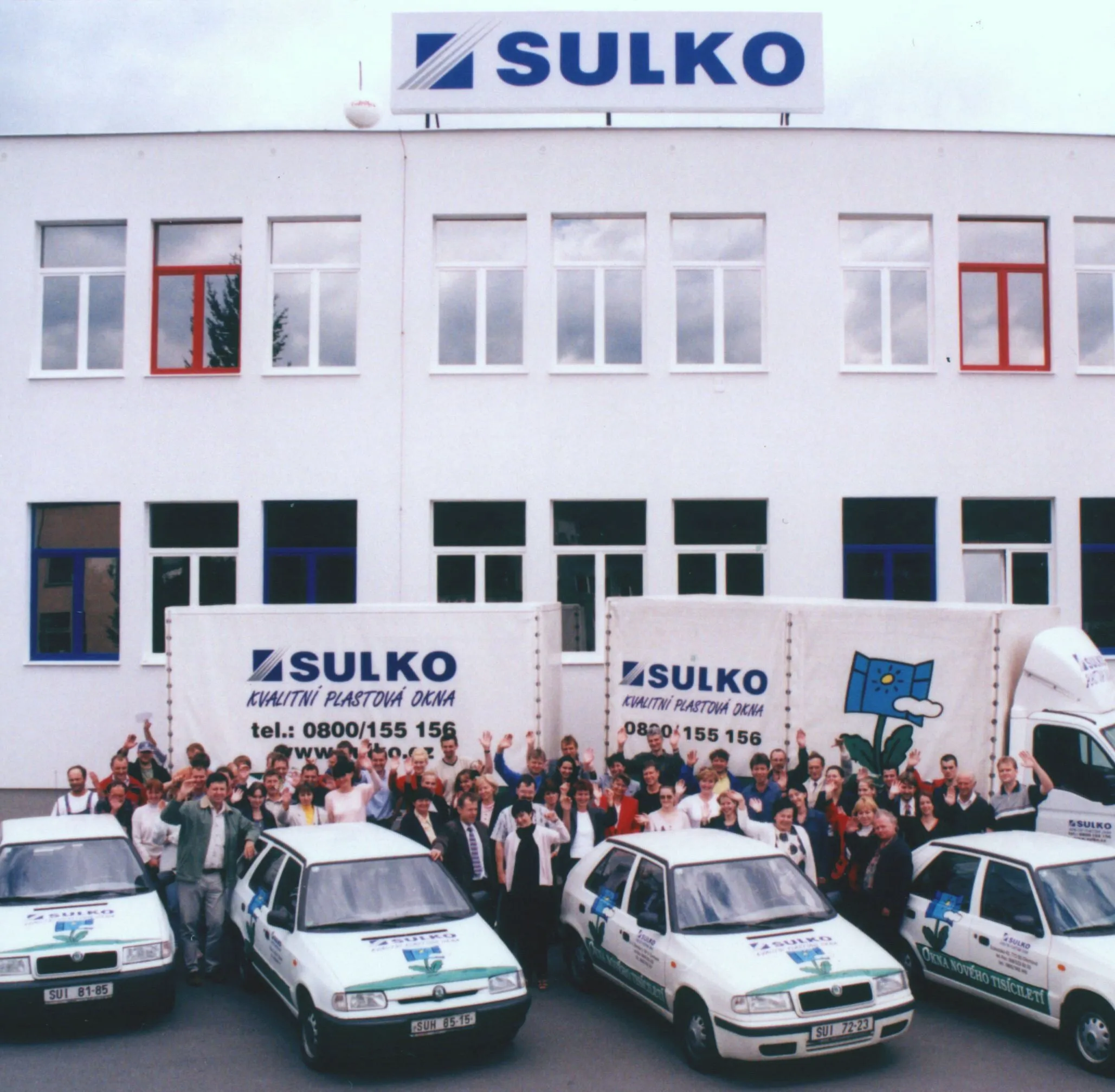 Skupina zaměstnanců stojící před budovou Sulko s firemními vozy a nákladními vozy s logem Sulko a nápisem kvalitní plastová okna.