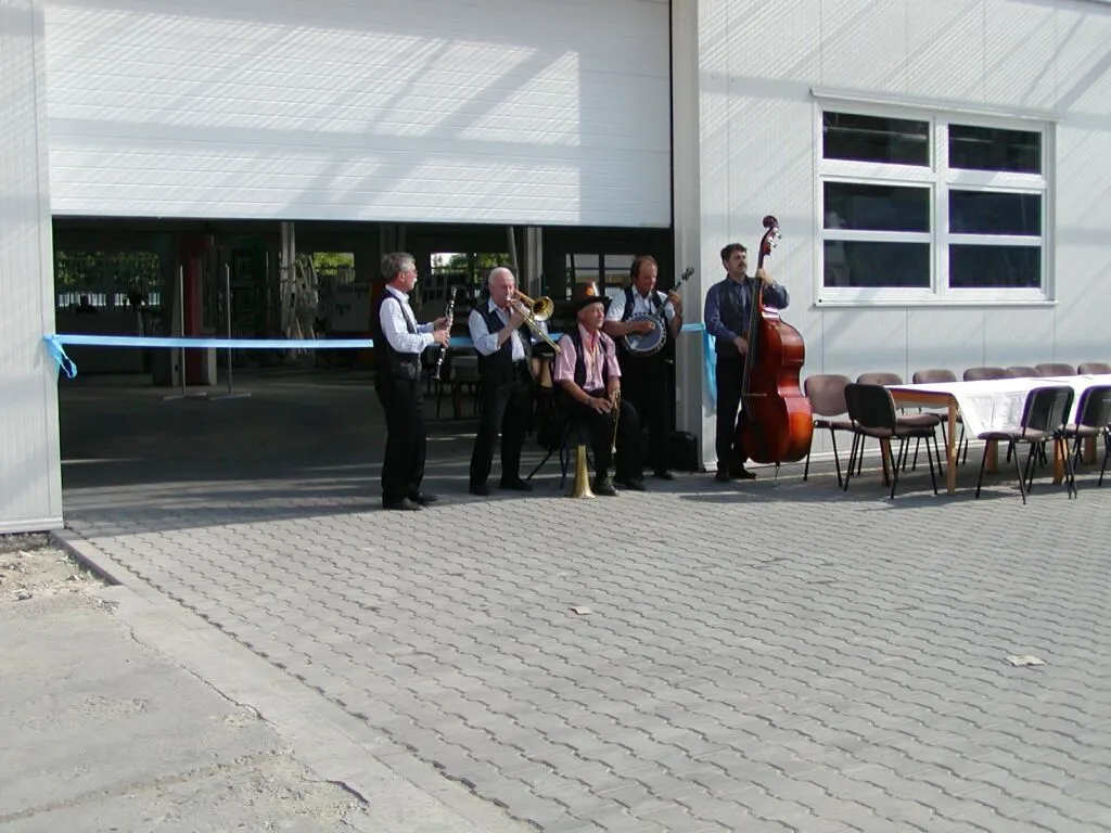 Skupina pěti hudebníků s dechovými a strunnými nástroji stojí před moderní budovou s otevřenou velkou branou a modrou stužkou, vedle nich jsou stoly a židle.