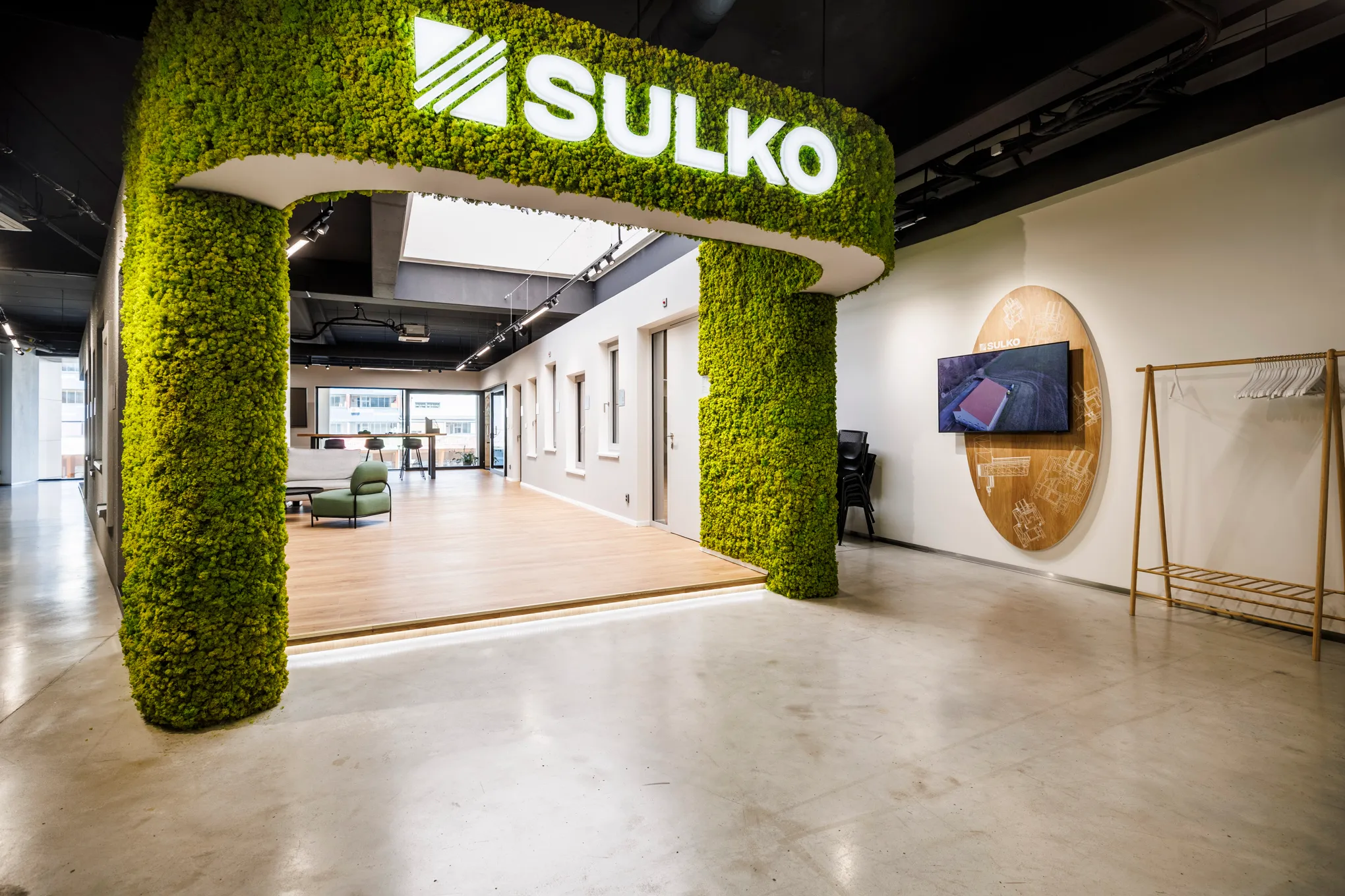 Modern office entrance with green moss-covered arch displaying illuminated SULKO logo, wooden floor area with seating, and TV mounted on the wall.