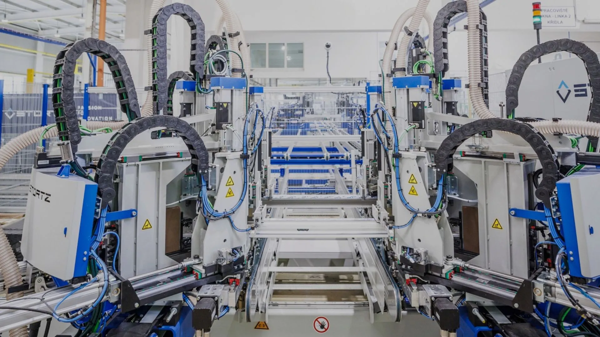 Průmyslový robotický stroj s modro-šedými součástmi a kabeláží v moderní výrobní hale.