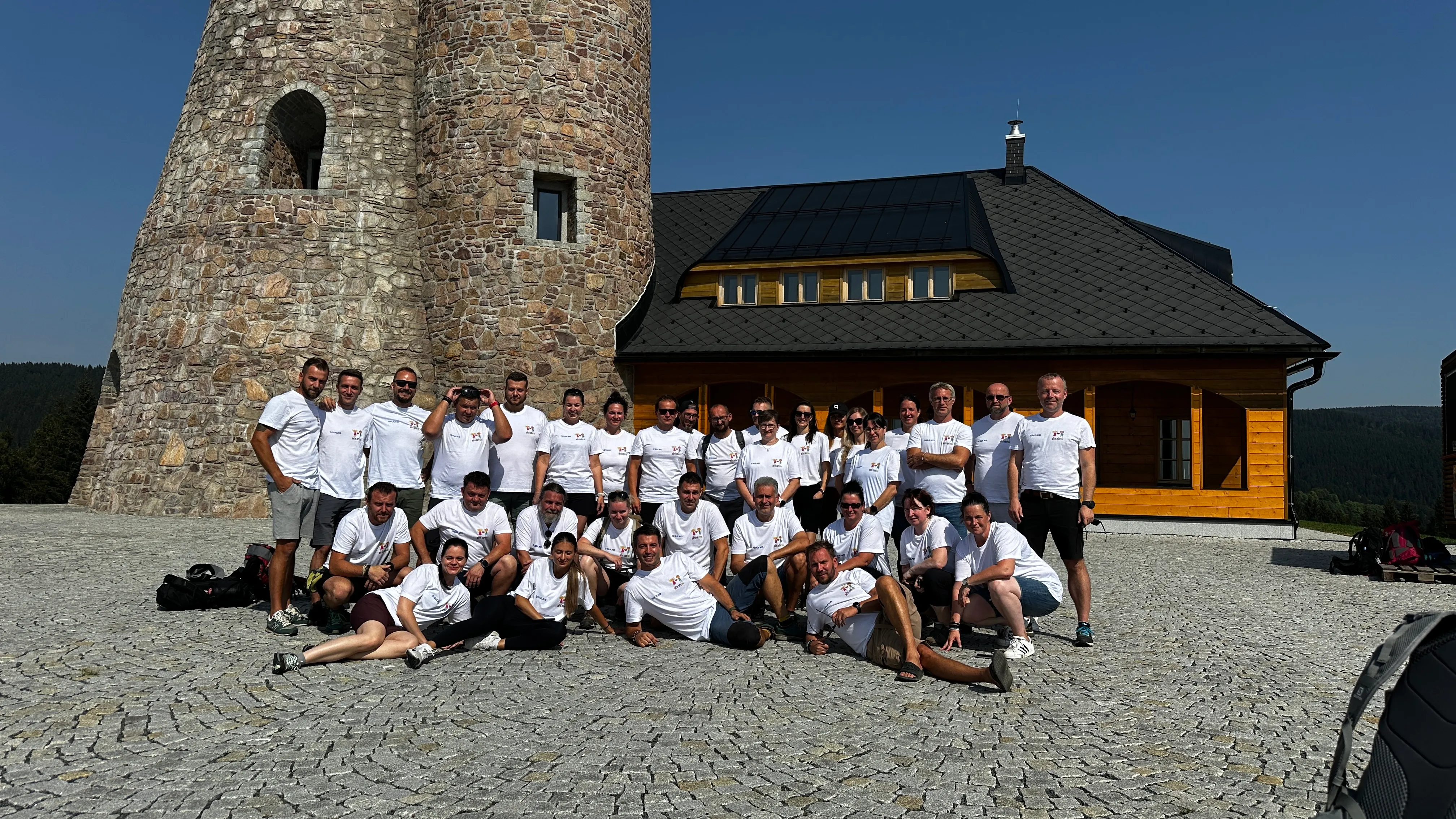 Skupina lidí v bílých tričkách pózuje před kameným věžovým objektem a dřevěnou chatou za slunného dne.