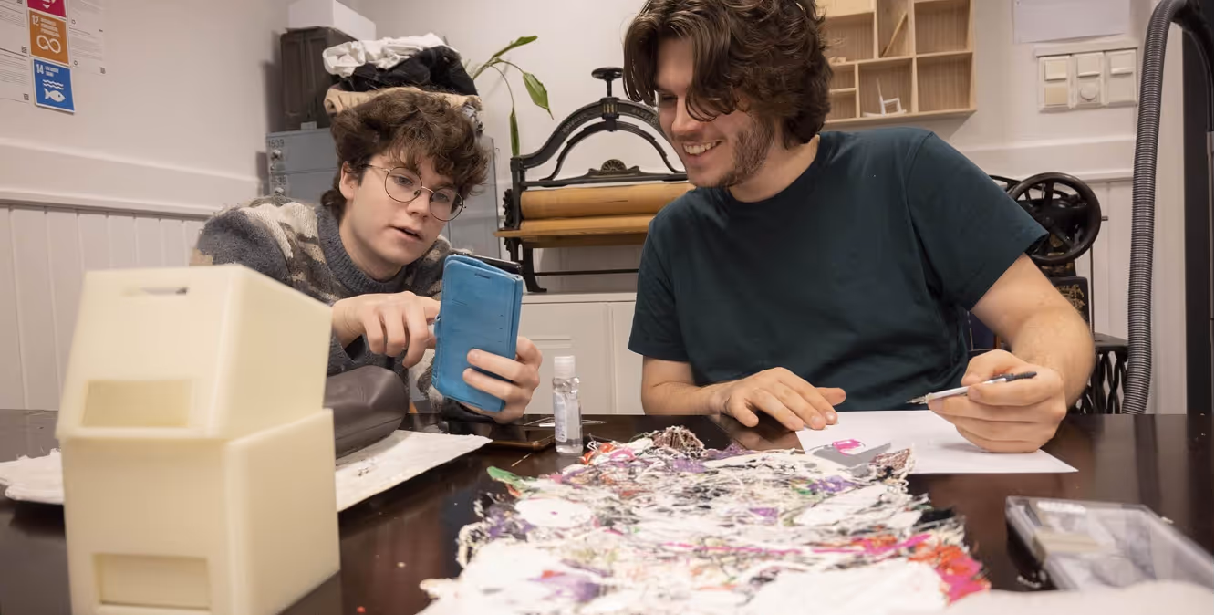 To unge personer samarbeider ved et bord med stoffprøver og verktøy i en kreativ arbeidsplass.