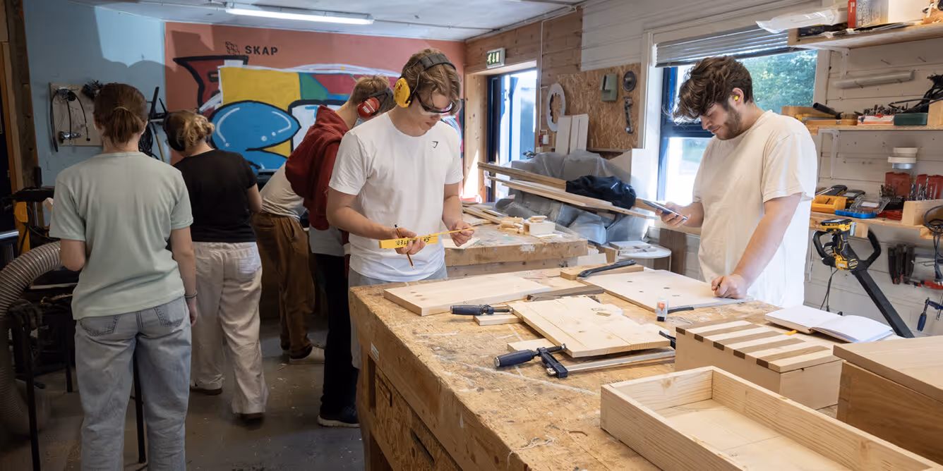 En gruppe unge mennesker jobber med trearbeid i et verksted, noen bruker hørselsvern og målebånd.