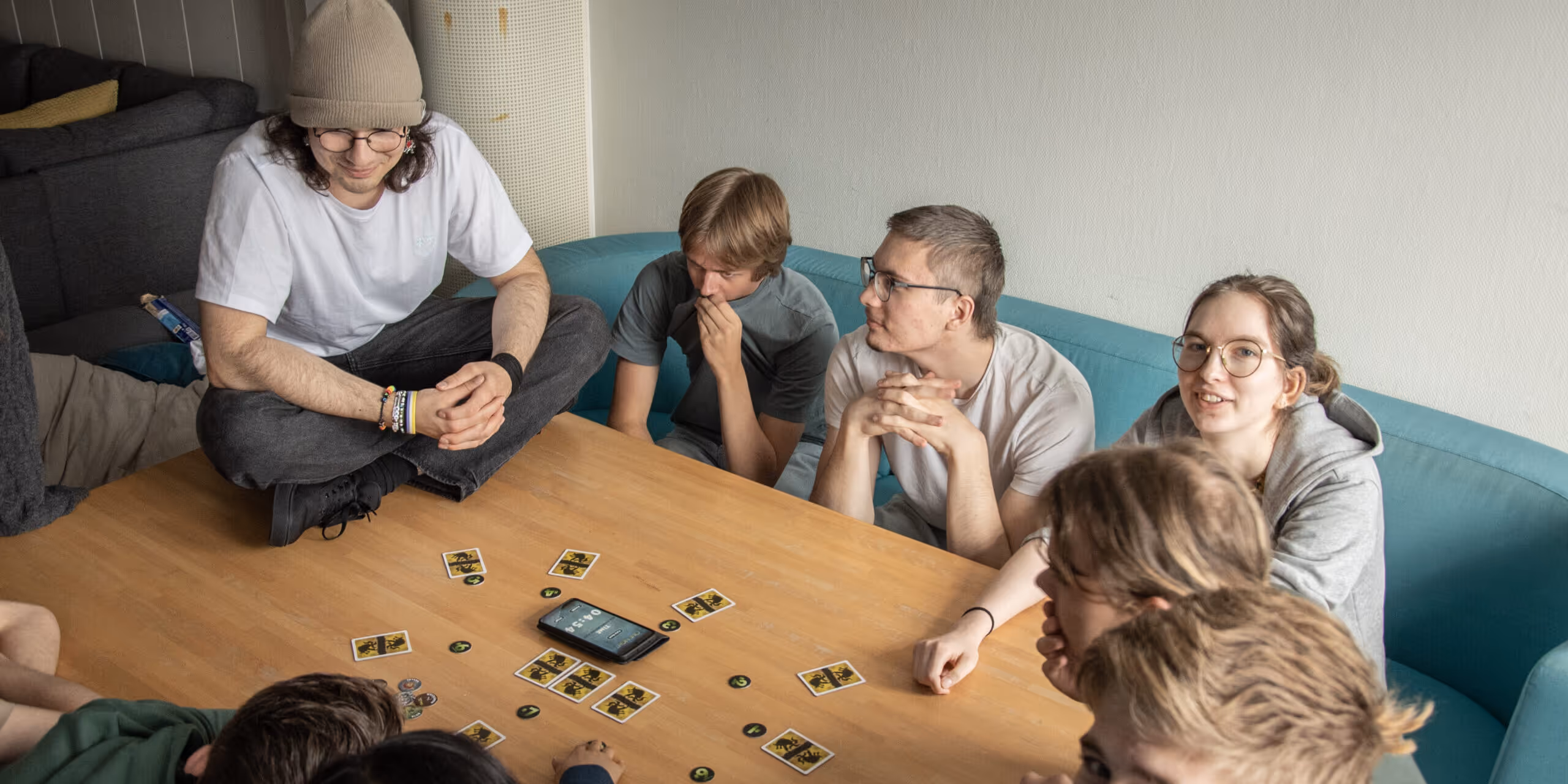 En gruppe unge sitter rundt et trebord og spiller et brettspill med kort og brikker, en smarttelefon er plassert midt på bordet.