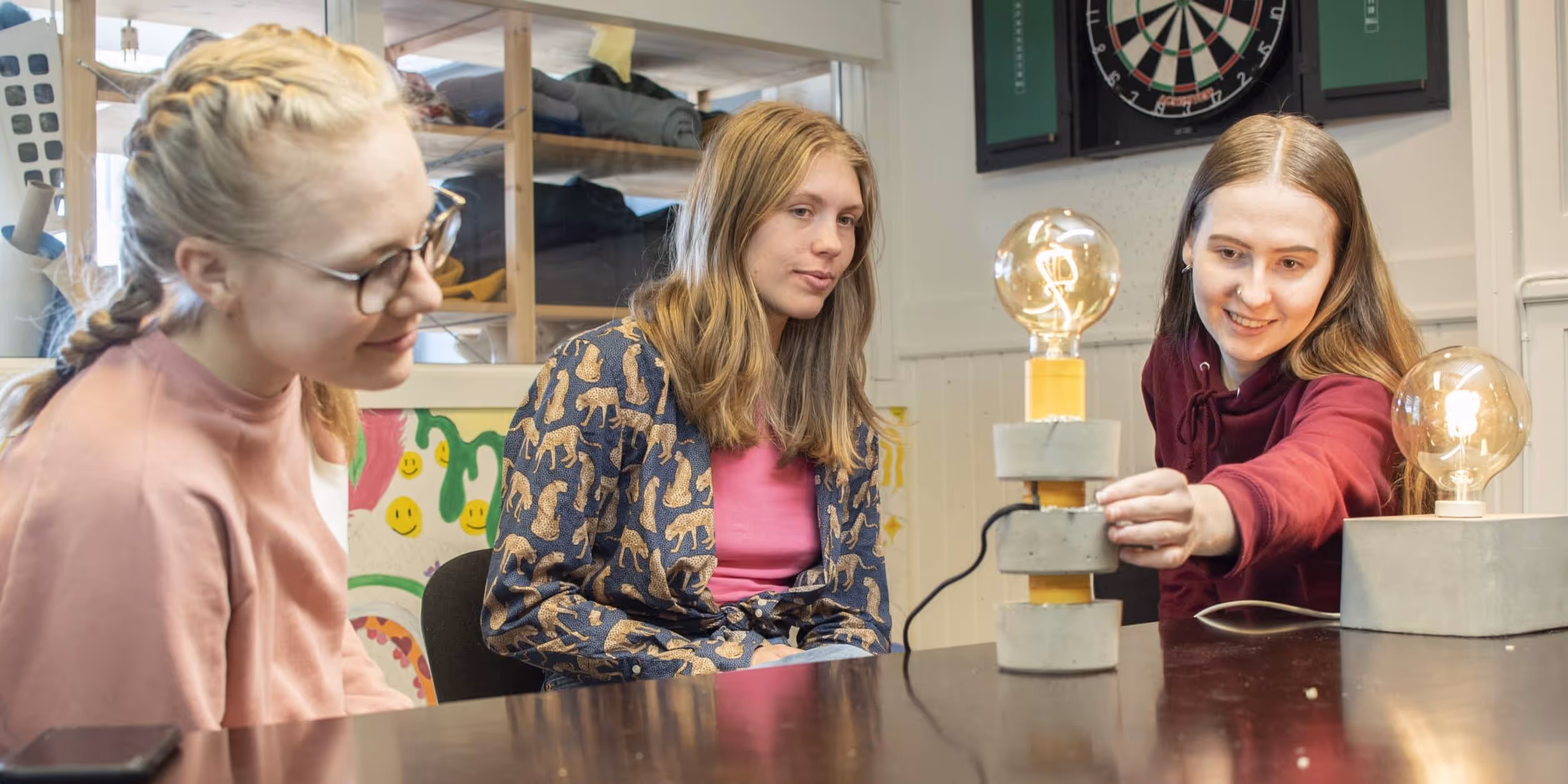 Tre unge kvinner sitter rundt et bord og ser på to unike bordlamper med synlige lyspærer.