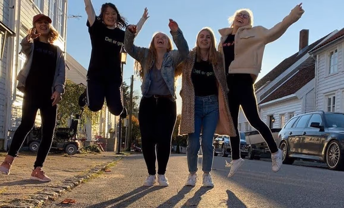 Fem unge kvinner ler og hopper i en solfylt gate med hvite treboder og parkerte biler i bakgrunnen.