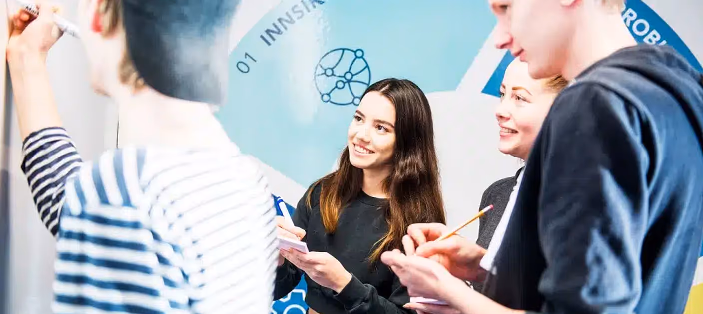 Gruppe ungdommer i et klasserom eller møtelokale skriver og diskuterer foran en tavle.