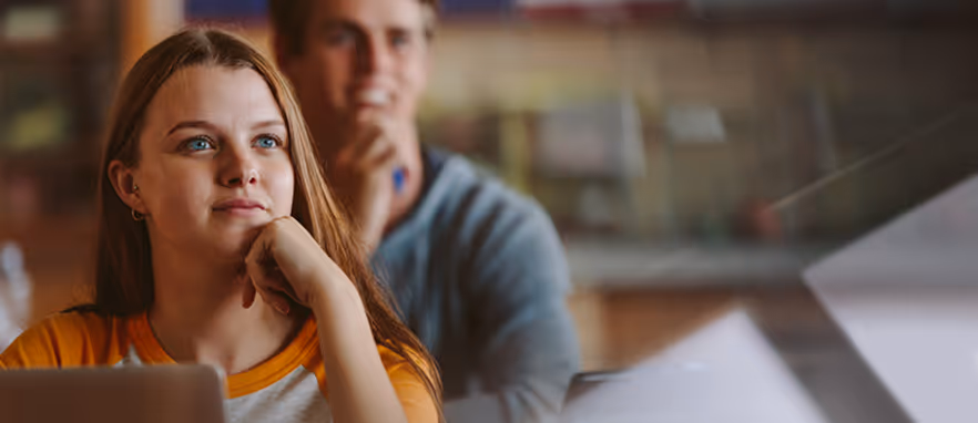Ung kvinne med langt hår som ser drømmende ut mens hun hviler hodet på hånden i et klasserom.