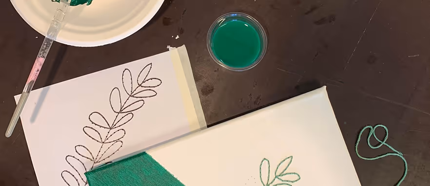 Malerutstyr med grønn maling, pensel, helleblad med bladtegning, og et lerret med delvis malt grønn trekant og broderi av blader.