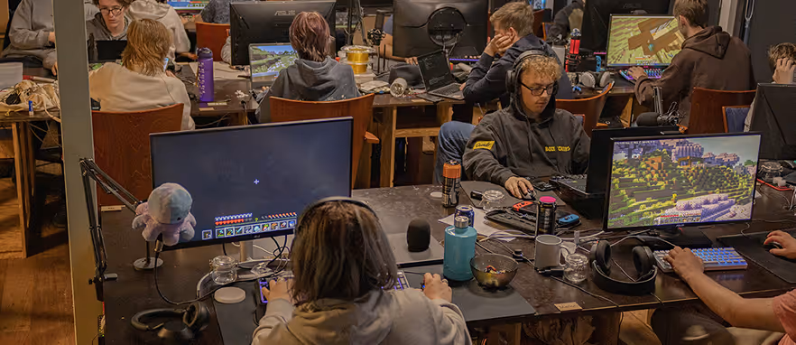 Personer i et rom som spiller og streamer Minecraft på datamaskiner med skjermer på et felles bord.