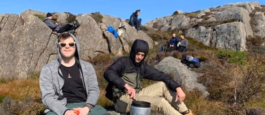 To unge menn sitter på steiner i et fjellandskap med sekker rundt seg under en klar blå himmel.