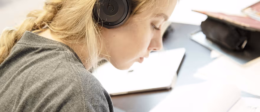 Ung kvinne med langt lyst hår og hodetelefoner som leser eller studerer ved et skrivebord med bøker.