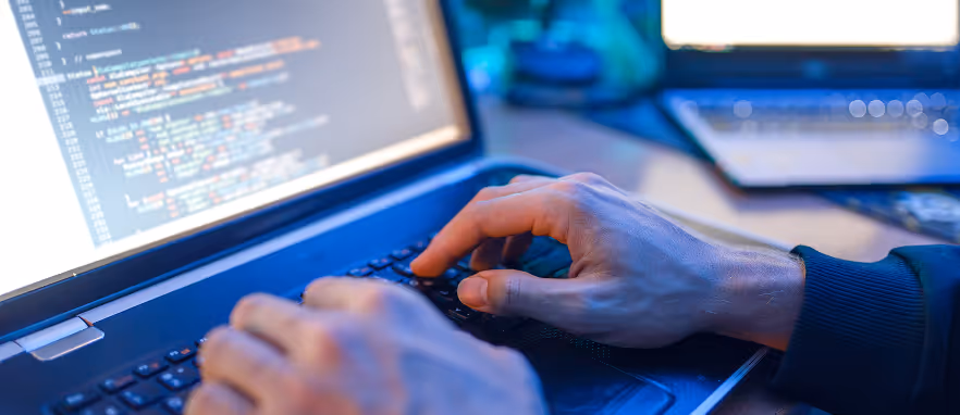 Hender som skriver kode på et tastatur foran en bærbar dataskjerm med kode.