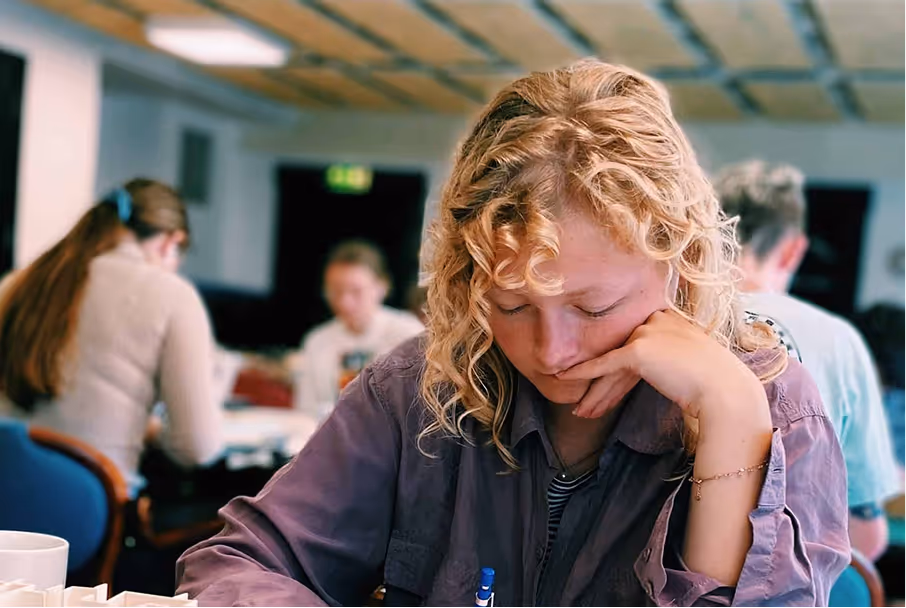 Ung kvinne med krøllete blondt hår som skriver mens hun hviler hodet på hånden i et klasserom eller bibliotek.