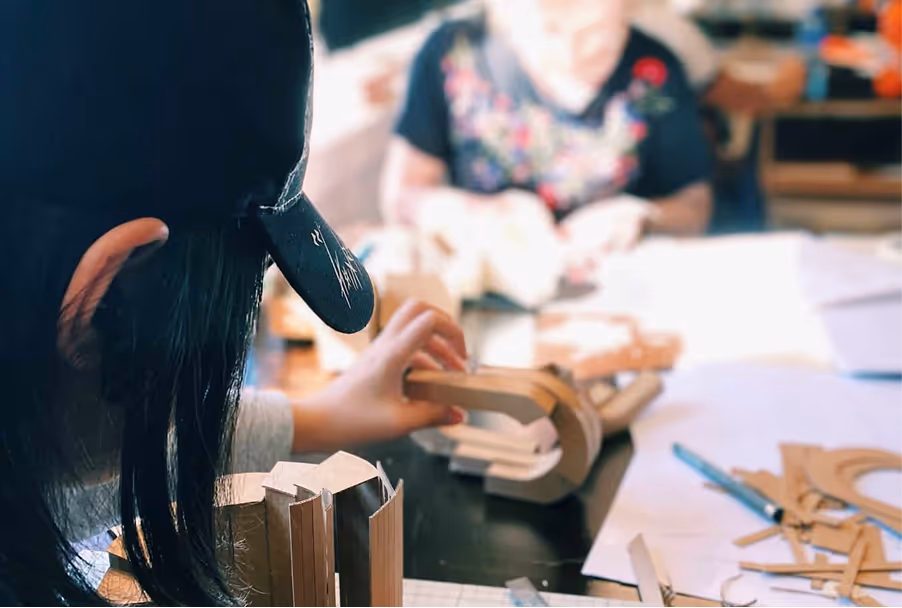 Person med svart caps håndterer små trebiter på et bord med en annen person i bakgrunnen.