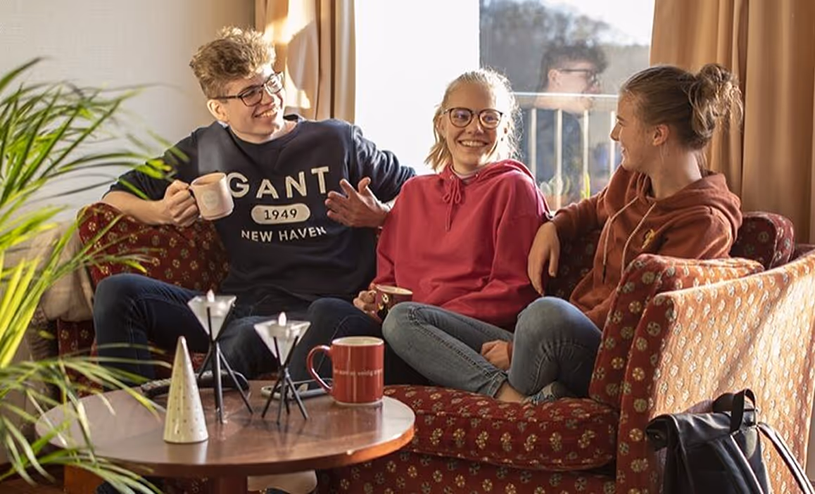 Tre unge mennesker sitter på en sofa og prater mens de drikker kaffe i et solfylt rom.