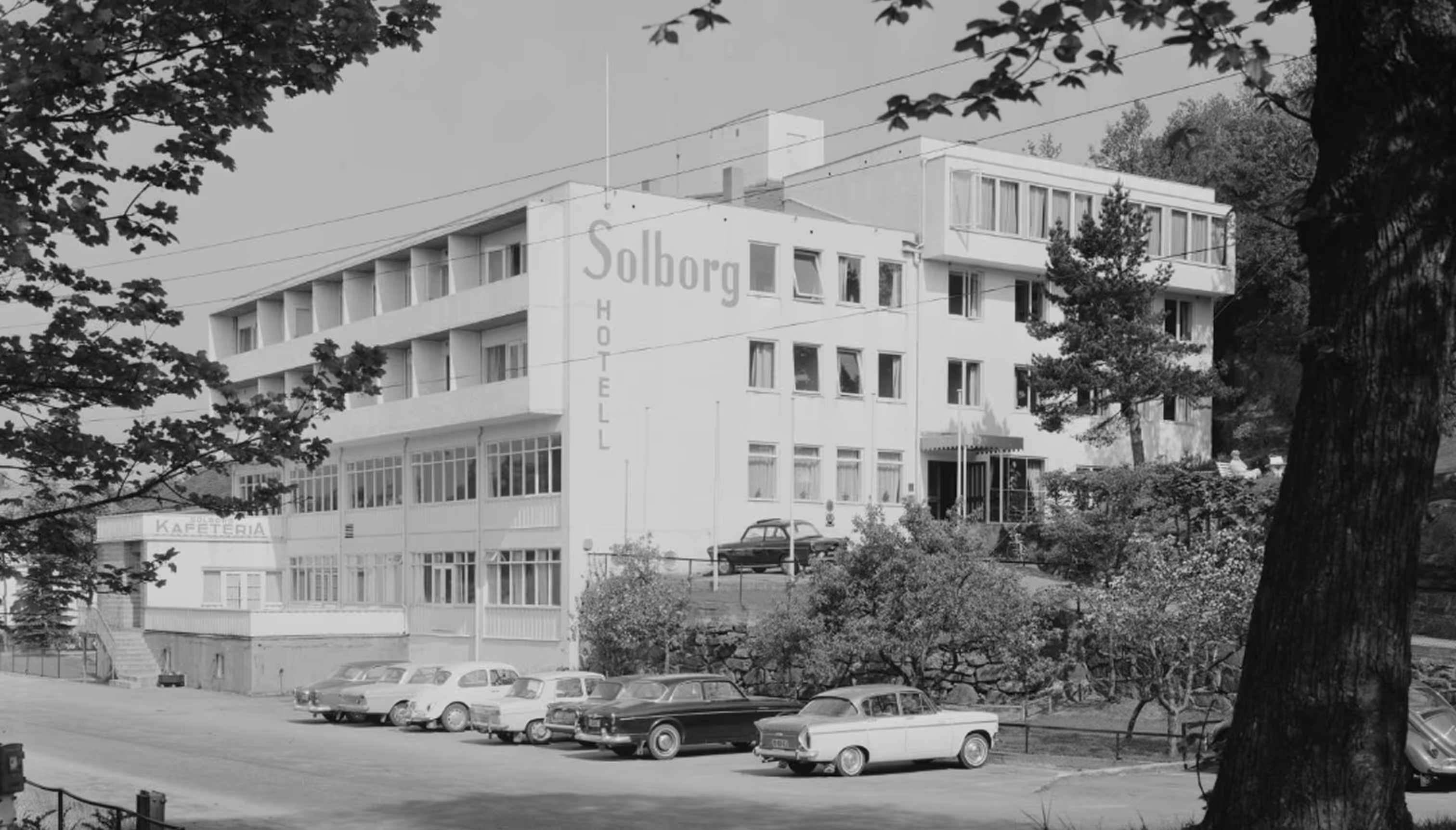 Svart-hvitt bilde av Solborg hotell med flere parkerte biler utenfor og omgitt av trær.