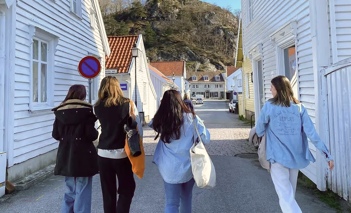 Fire unge kvinner med vesker går ned en gate med hvite trehus og et fjell i bakgrunnen under blå himmel.