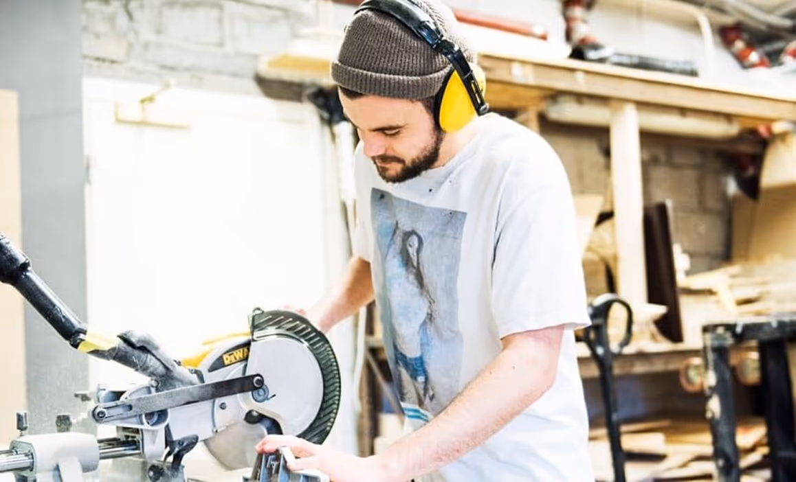 Mann med lue og gule hørselsvern bruker en kapp-/gjæringssag i et verksted.