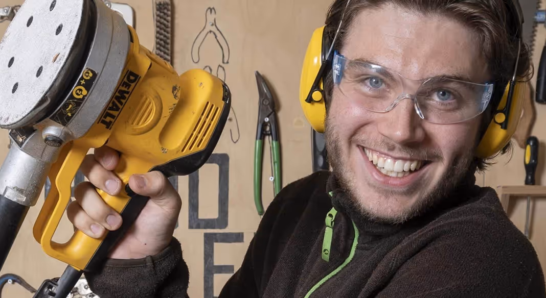Smilende mann med vernebriller og hørselsvern holder en gul håndholdt DeWalt slipemaskin i et verksted.