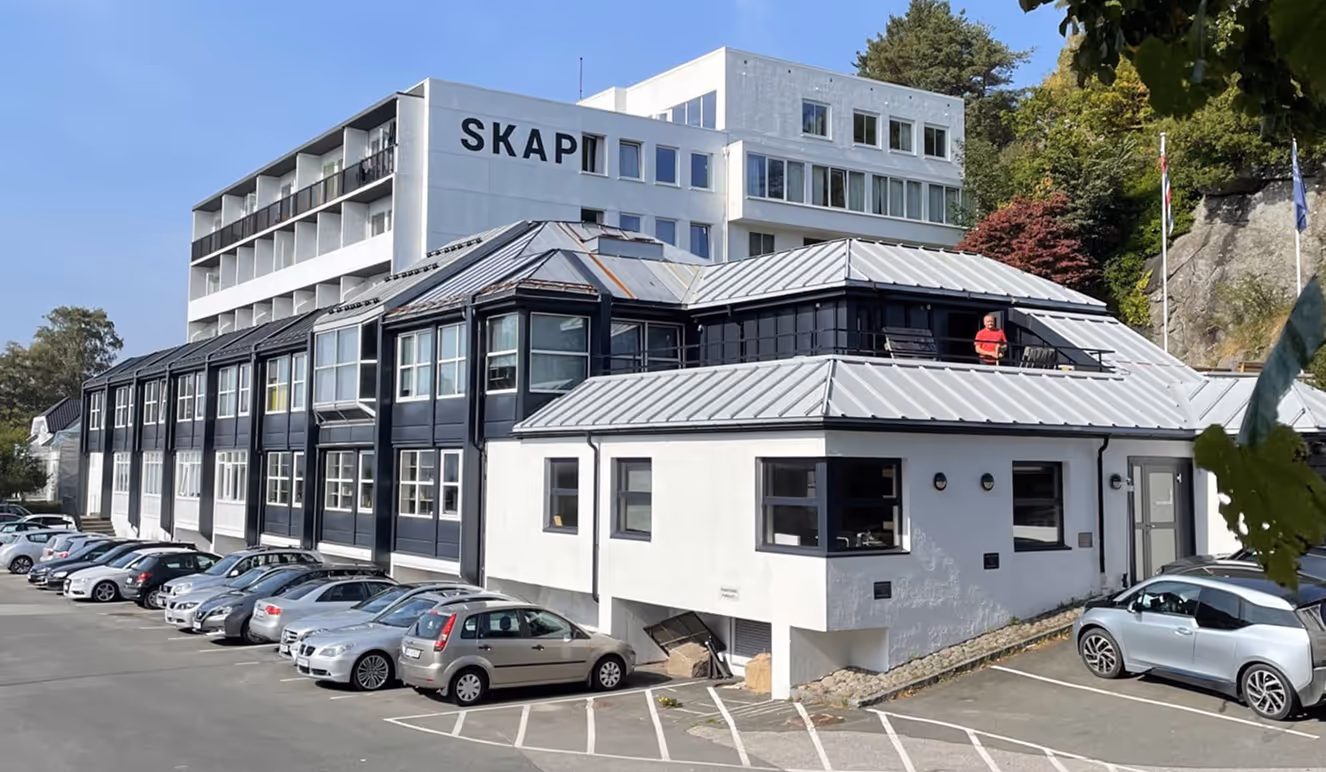 Bygning med skiltet SKAP og flere parkerte biler på en solrik dag.