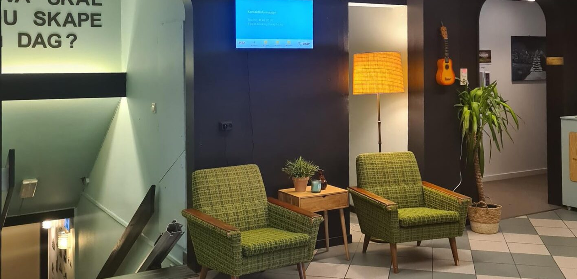 To grønne lenestoler med trebord i mellom, gulvlampe, inneplante i kurv, og en liten gitar hengt på veggen i et koselig rom.