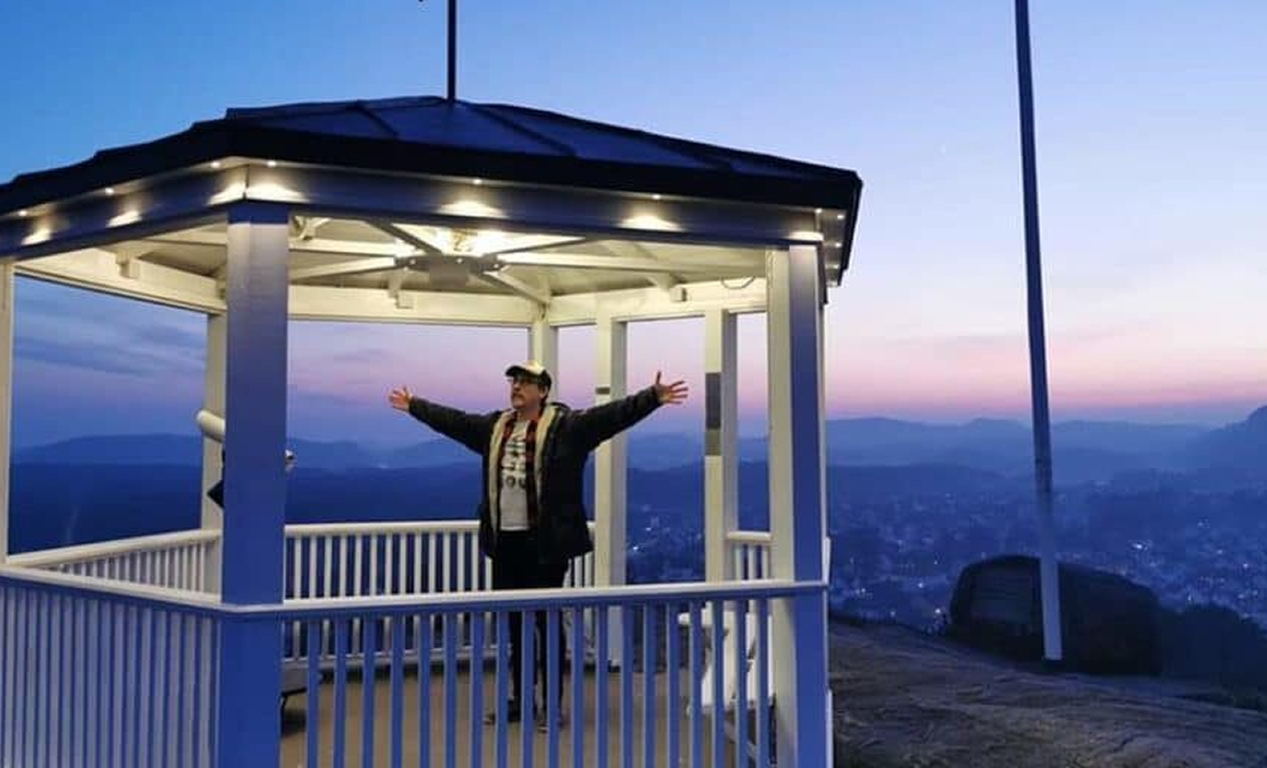 Person står med armene åpne i en opplyst åpen hagestue med utsikt over en by og fjell under solnedgang.