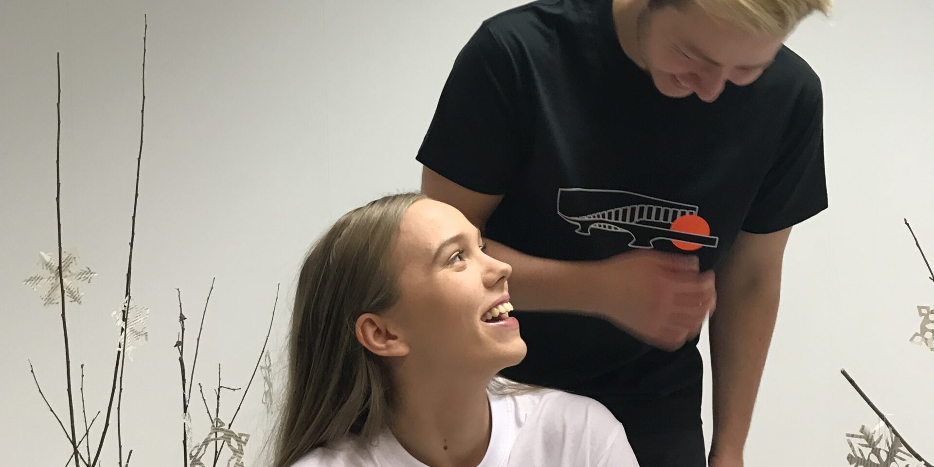 Ung kvinne ser opp og smiler mens en ung mann ler ved siden av henne, begge i et rom med hvit vegg og dekorative kvister med papirstjerner.