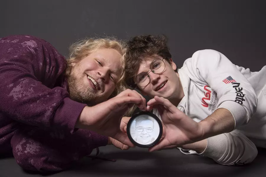 To unge menn ligger ned og danner et hjerte med hendene rundt en forstørrelsesglass som viser et ansikt.