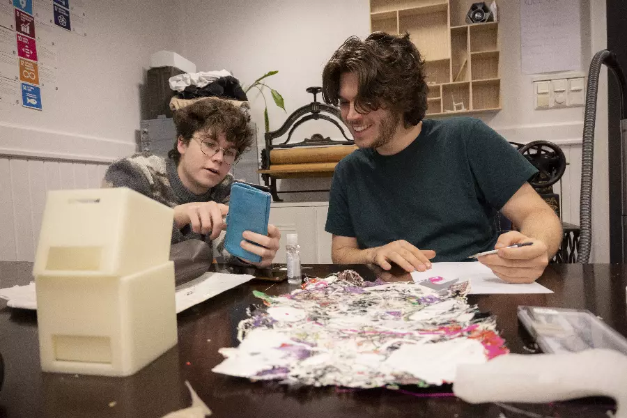 To unge menn sitter ved et bord med kunstprosjekt og ser på noe på en mobiltelefon, omgitt av kreative materialer og verktøy.
