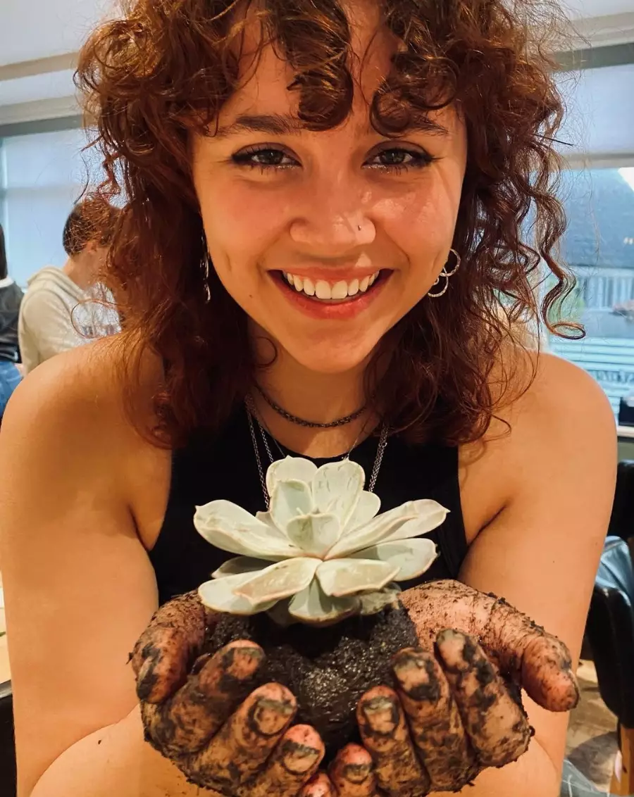 Smilende kvinne med krøllete hår som holder en hvit sukkulentplante med jord på hendene.