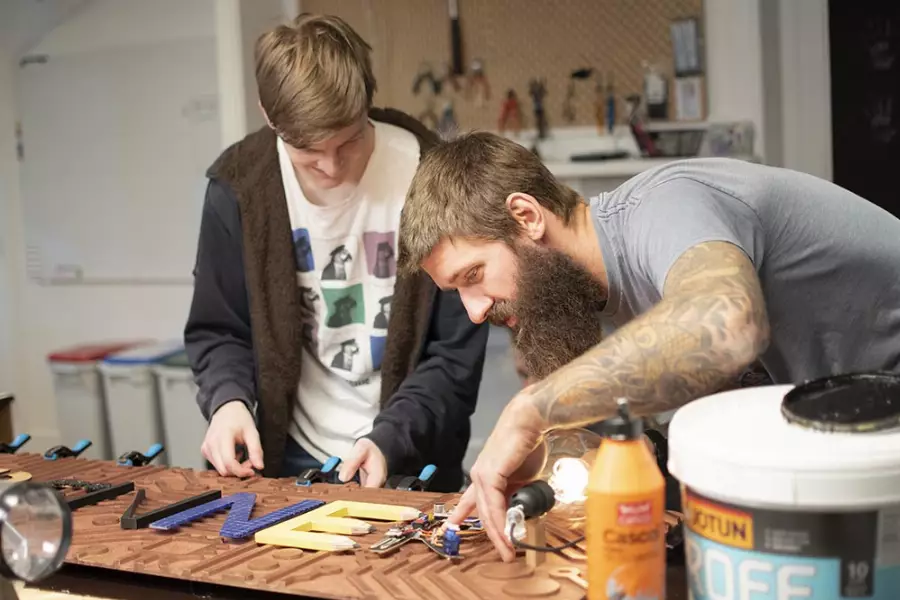 To menn som samarbeider om et treprosjekt på arbeidsbenk, en med skjegg og tatoveringer og en yngre mann som ser ned.