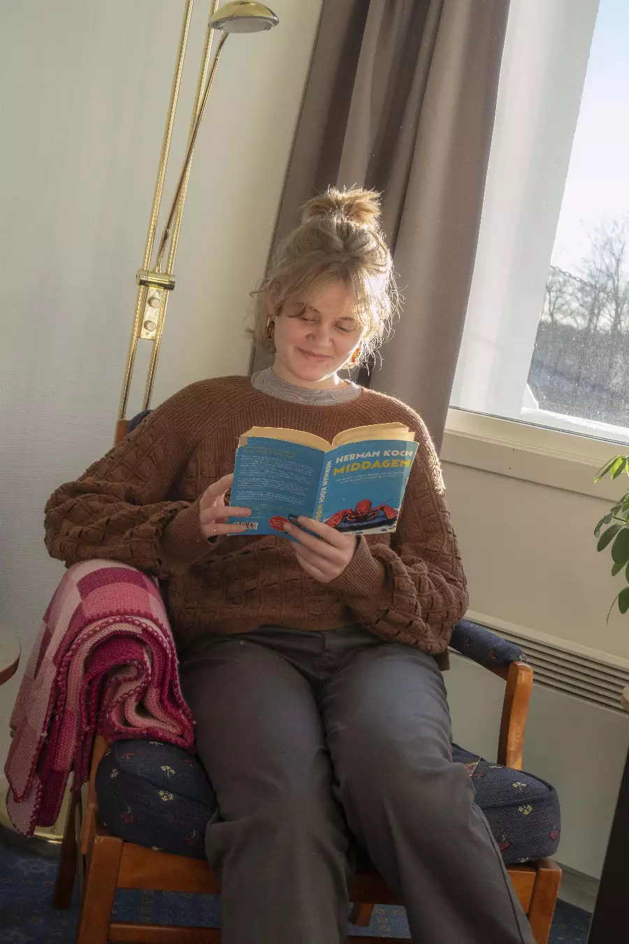 Ung kvinne med brunt flettet skjerf leser bok mens hun sitter i en stol ved et vindu med gardiner.