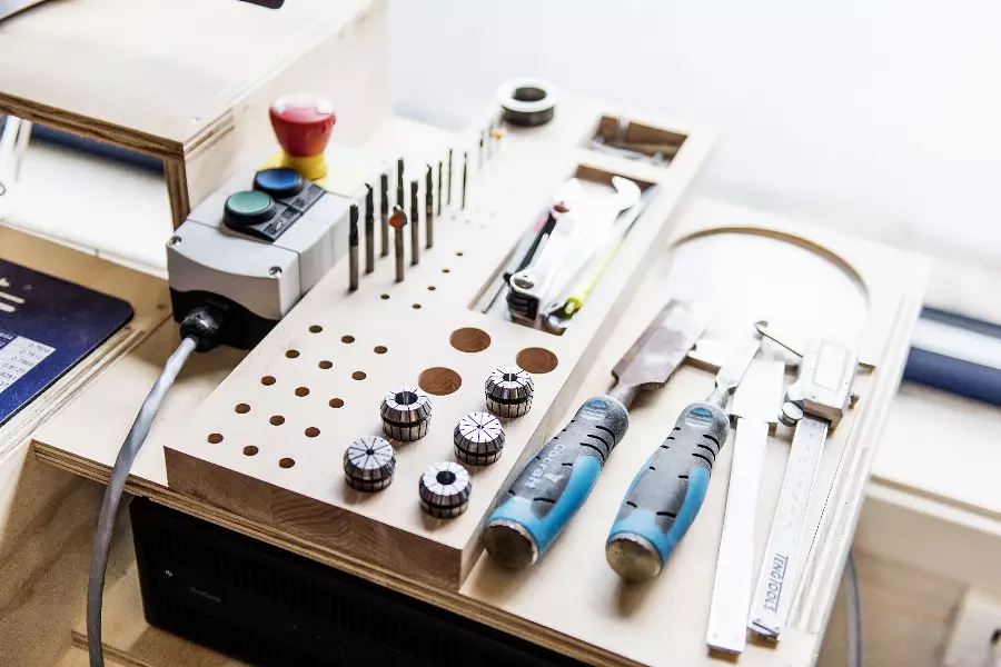 Verktøystativ med borefrasker, håndverktøy med blå og svarte håndtak, samt et måleverktøy plassert på et lyst trebord.