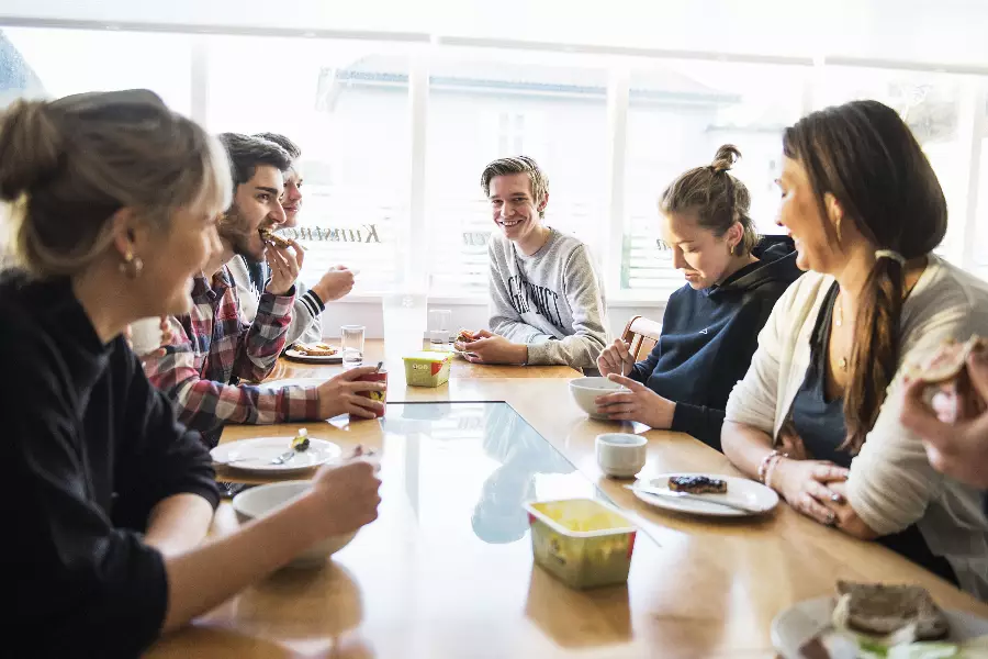 En gruppe unge mennesker som sitter rundt et bord og spiser sammen, noen smiler og andre spiser.