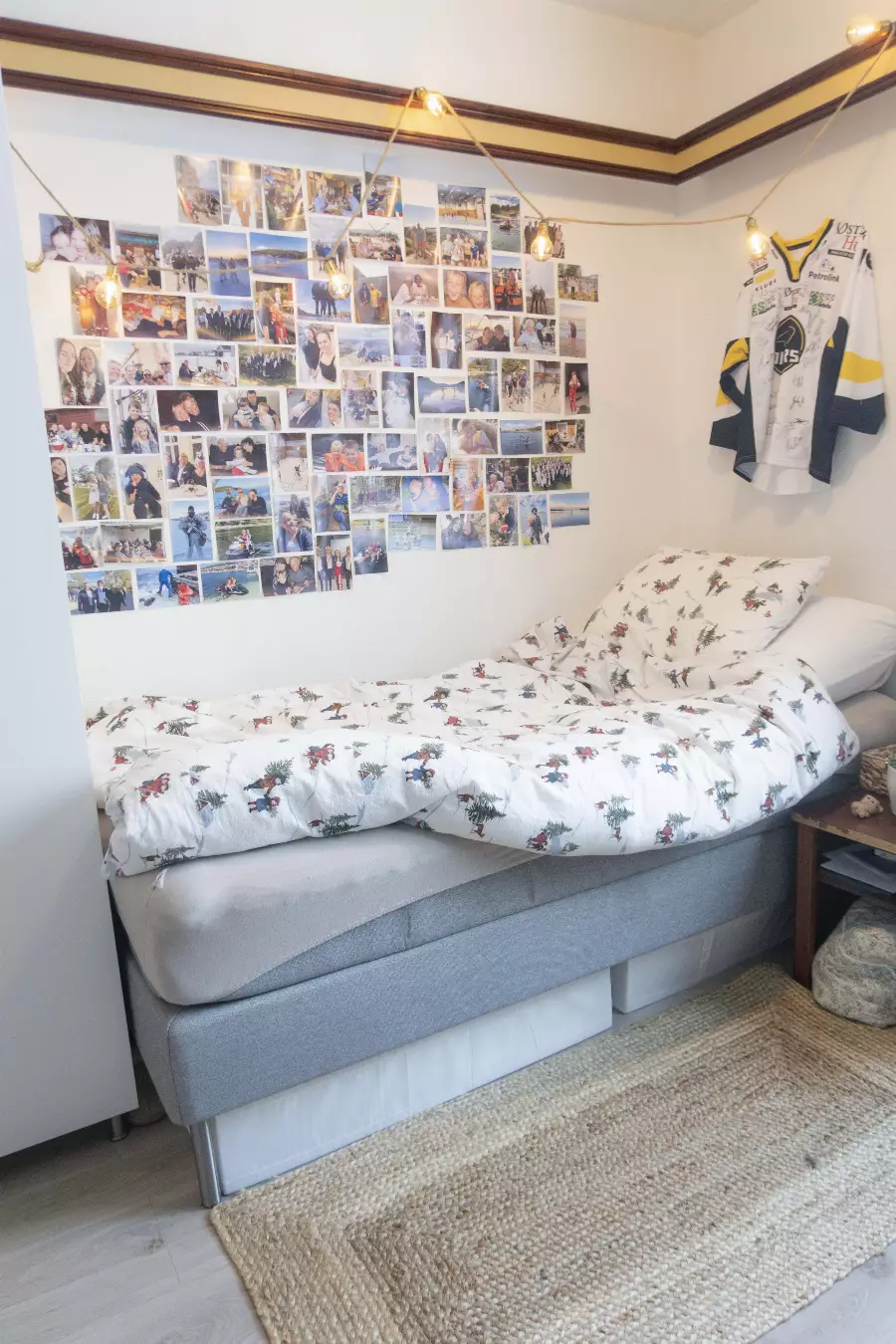 Soverom med enkeltseng med juletema sengetøy, vegg dekorert med familiebilder og en signert hockeytrøye, opplyst av hengende lyspærer.