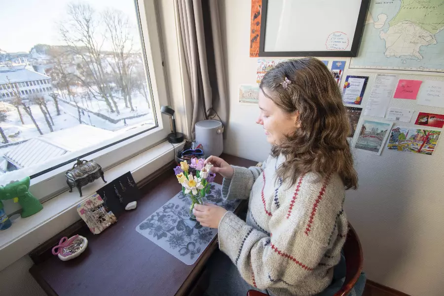 Kvinne sitter ved et skrivebord ved et vindu og ordner en bukett med fargerike blomster innendørs.