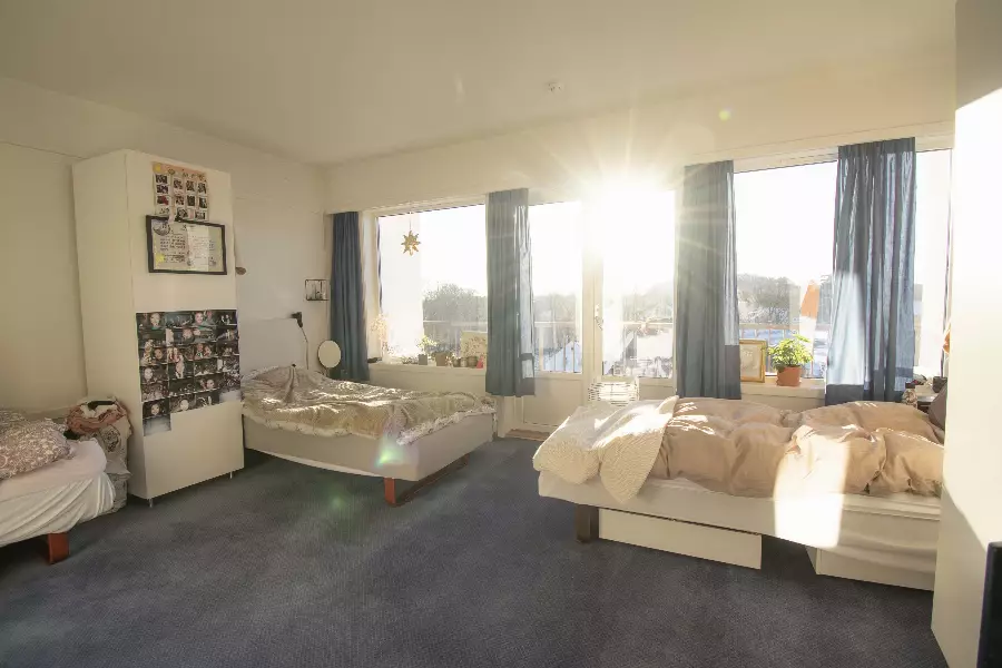 Solsidig soverom med to dobbeltsenger, blå tepper, blå gardiner og store vinduer som slipper inn mye dagslys.