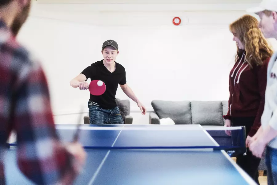 En gruppe unge spiller bordtennis innendørs, en gutt med svart t-skjorte og caps slår ballen over bordet.