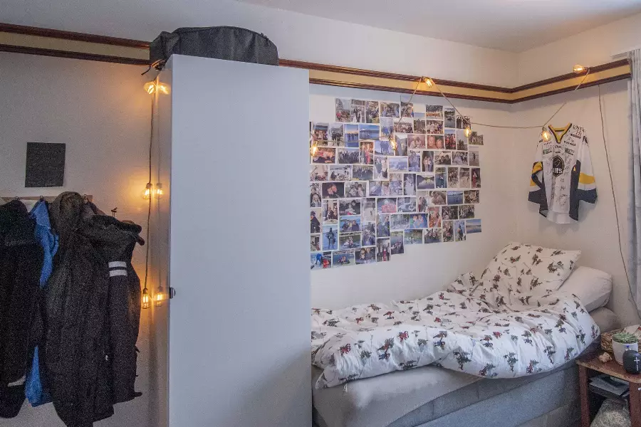 Soverom med seng og sengetøy med mønster, veggdekorasjon av mange små bilder, hengt opp sportsdrakt, og en garderobe med jakker hengende ved siden av.