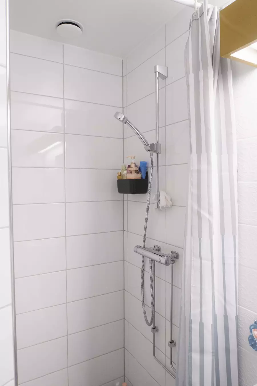 Dusj med hvite fliser, krom hånddusj, sort hylle med såpeflasker, og et hvitt og gråstripet dusjforheng.
