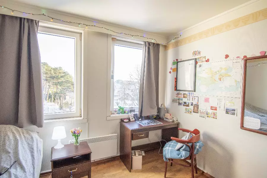 Lyst rom med to vinduer, et skrivebord med stol dekket med et teppe, nattbord med lampe og blomster, og vegg dekorert med kart og postkort.