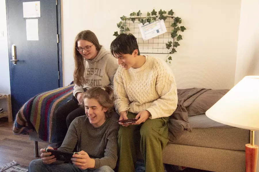 Tre ungdommer sitter sammen i et rom og spiller videospill mens de ler.