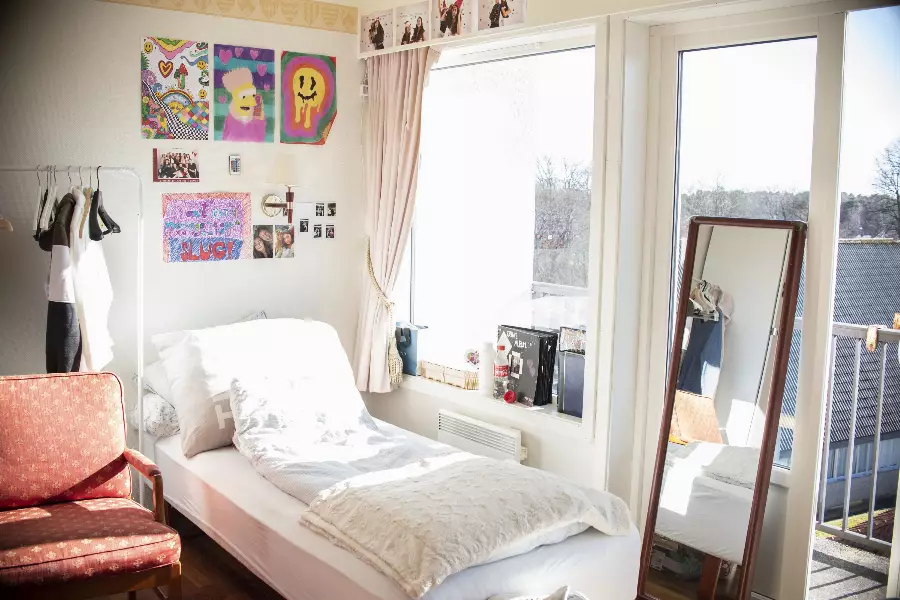 Lyst soverom med enkeltseng, rød stol, fargerike plakater på veggen og stort vindu med utsikt mot balkong.