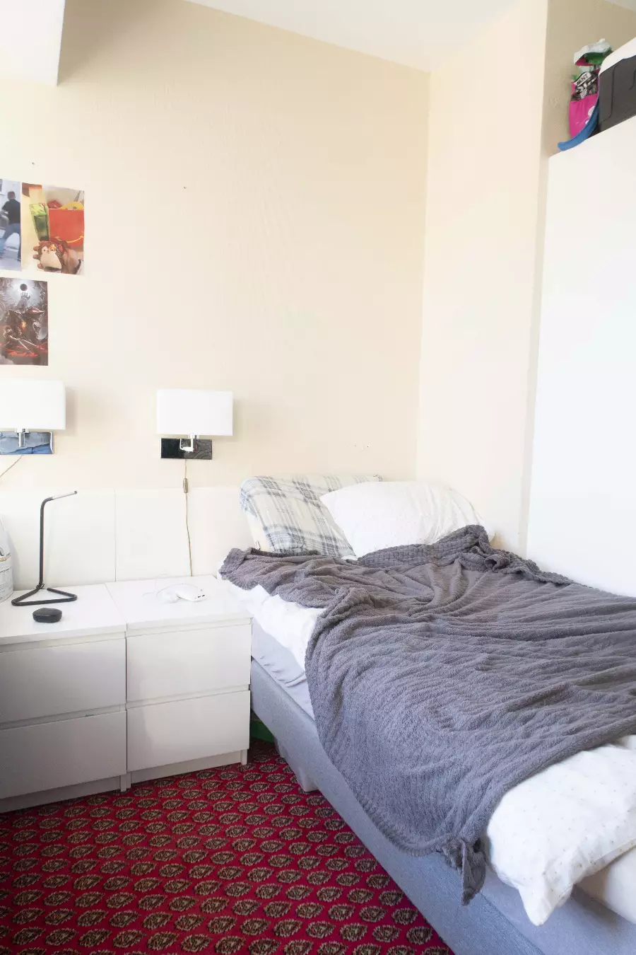 Soverom med enkeltseng dekket med grått teppe, hvite puter, hvitt nattbord med bordlampe, og rødt mønstret teppe på gulvet.